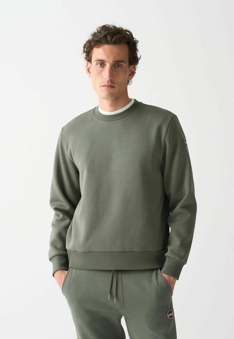 Hanorac verde din material moale, cu guler rotund, mansete și tiv cu dungi, având un patch cu logo pe manșa stângă. La set cu pantaloni asortați.