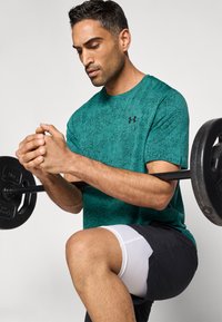 Γαλάζιο αθλητικό μπλουζάκι με υφή και κοντά μανίκια, με μαύρο λογότυπο Under Armour, σε συνδυασμό με μαύρο σορτς και λευκά συμπιεστικά στρώματα, σηκώνει βάρη.