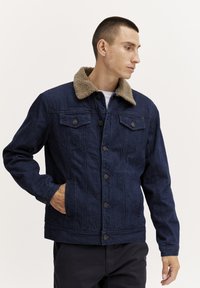 Donkerblauwe denim jas met een beige nepbont kraag. Bevat twee borstzakken en twee zijzakken, met metalen knopen.