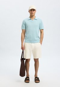 Polo shirt tricoté bleu clair à manches courtes, shorts crème, sac fourre-tout marron et sandales foncées sur un fond neutre.