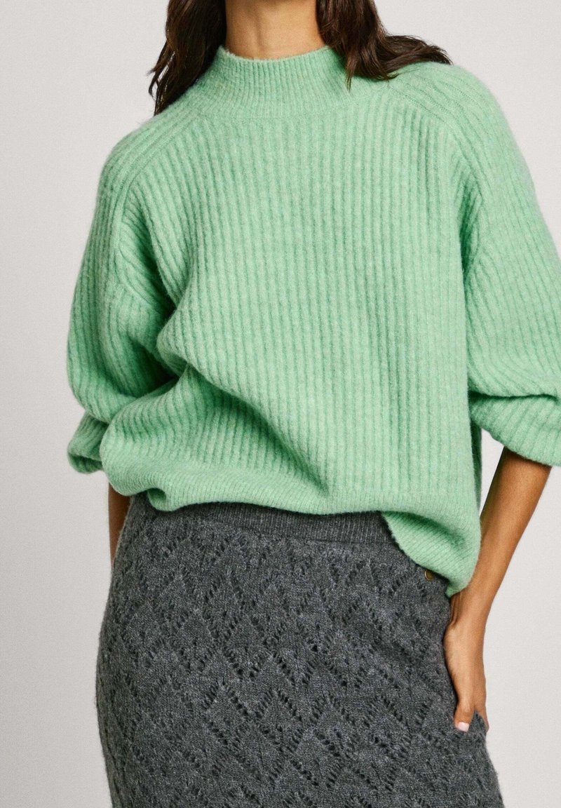 Mintgrüner, gerippter Pullover mit hohem Kragen und oversized Schnitt, kombiniert mit einem strukturierten grauen Rock mit Rautenmuster.