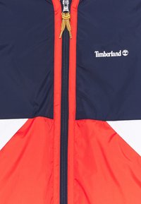 Rote, marineblaue und weiße Windbreaker-Jacke mit einem vorderen Reißverschluss und einem goldfarbenen Zug, sowie einem Timberland-Logo auf der Brust.