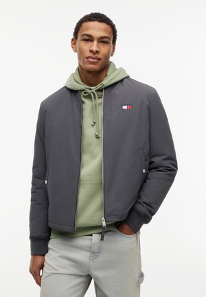 Άνδρας που φοράει ανοιχτό πράσινο φούτερ με κουκούλα κάτω από σκούρο γκρι φερμουάρ μπουφάν, με ανοιχτό γκρι τζιν, με τα χέρια εν μέρει στις τσέπες, στέκεται μπροστά σε λευκό φόντο.