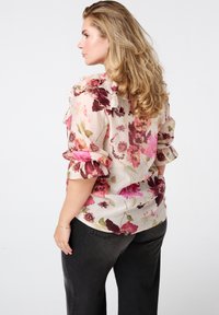 Bloemrijke blouse met ruches aan de mouwen in tinten van roze, rood en groen op een lichte achtergrond; gecombineerd met zwarte, wijde broeken.