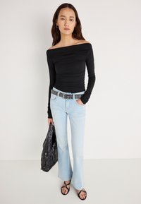 Zwarte off-shoulder top met lange mouwen, lichtblauwe wijde spijkerbroek, met studs versierde riem, zwarte gestructureerde tas, zwarte platte sandalen, model staand tegen een effen achtergrond.