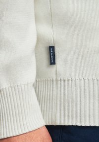 Gros plan sur le poignet et l'ourlet d'un pull blanc en maille, avec une étiquette en tissu "MR MARVIS" visible sur la couture latérale.