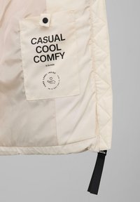 Beżowa pikowana tkanina z prostokątną kieszenią oznaczoną napisem "CASUAL COOL COMFY", z zatrzaskiem i czarnym materiałowym paskiem wiszącym poniżej.