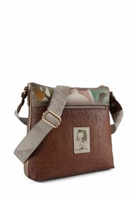 Braune, strukturierte Leder-Umhängetasche mit gemustertem Deckel, verstellbarem Riemen und einem dekorativen Anhänger mit einer Charakterillustration.