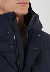 Giacca imbottita blu navy con colletto alto, dotata di chiusura con zip, bottoni a pressione e dettaglio in rilievo "ANIMAL FREE" lungo la zip.