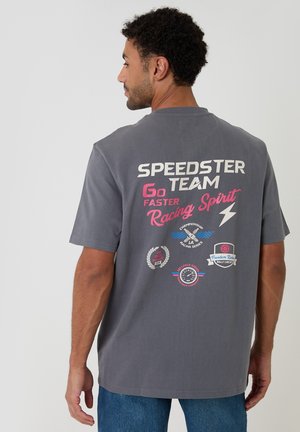 Mand set bagfra iført en grå T-shirt med farverig racertema tekst og grafik på bagsiden.