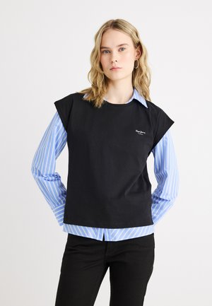 Žena s vlnitými blond vlasmi, oblečená v čiernom tielku cez modro-bielu pruhovanú košeľu s gombíkmi, stojaca so založenými rukami za chrbtom.