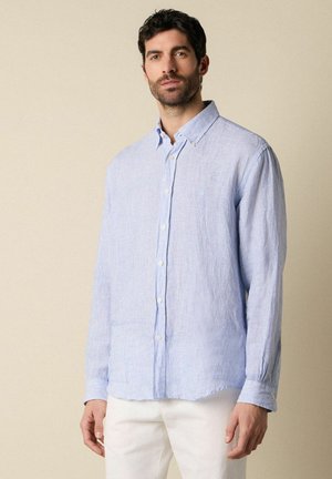 Mężczyzna stojący w jasnoniebieskiej, w paski, koszuli na guziki i białych spodniach na jednolitym beżowym tle.