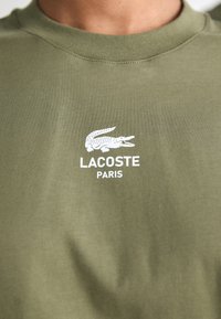 Žalia marškinėliai su balta Lacoste krokodilo logotipu ir tekstu "LACOSTE PARIS" ant krūtinės, arti išpjovos ir viršutinės liemens dalies.