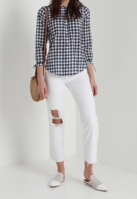 Camisa de botones en cuadros azul marino y blanco, combinada con jeans blancos desgastados. Un bolso de tejido y unas zapatillas slip-on blancas perforadas completan el look.