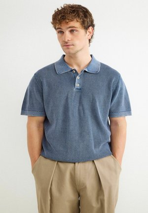 Młody mężczyzna z kręconymi włosami, ubrany w niebieską dzianinową koszulkę polo z krótkim rękawem i beżowe spodnie, ręce w kieszeniach, delikatnie spogląda na bok.