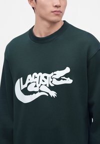 Homme portant un sweat-shirt vert foncé avec un motif crocodile stylisé blanc et le texte "Lacoste" sur la poitrine, faisant face légèrement à droite.