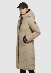 Beige isoleret lang jakke med hætte, quiltet design, sort lynlås, side lommer og en glat tekstur. Bæres med en sort kasket.