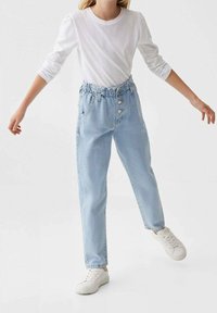 Pantalones de mezclilla azul claro con un corte relajado, cinturilla elástica y tres botones blancos. Combinados con una camisa blanca de manga larga y zapatillas blancas.