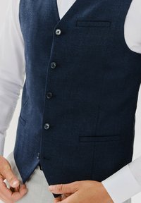 Granatowa kamizelka w jodełkę z pięcioma guzikami, dwiema kieszeniami i ostrymi klapami. Gładka tekstura, dopasowany krój, noszona na białej koszuli.