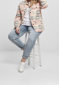 Patterned denim jacket in pink, grey, and white; white T-shirt; light blue rolled-cuff jeans; white sneakers; sitting on a white stool.