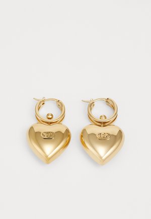 Paar glanzende gouden creooloorbellen met grote hartvormige hangers, gegraveerd met een klein logo op een effen witte achtergrond.