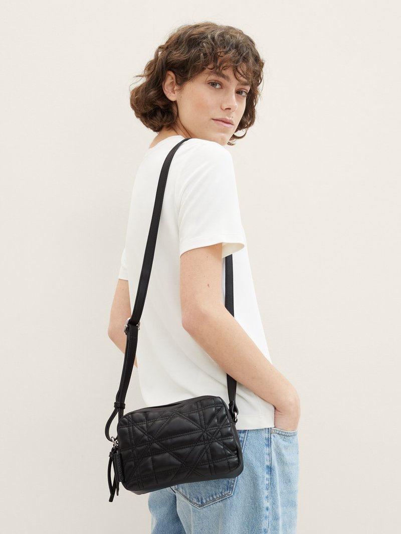 Femme aux cheveux courts et bouclés portant un T-shirt blanc et un jean, portant un sac bandoulière noir matelassé avec une sangle réglable sur l'épaule.