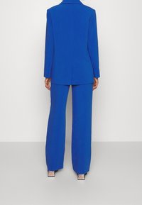 Blauw op maat gemaakt jasje en wijde broek. Glad stof, gestructureerd ontwerp, met een enkele split aan de achterkant en glanzende hakken.