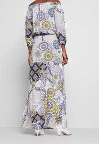 Robe longue épaules dénudées en tissu léger, présentant un motif floral et géométrique vibrant en bleu, jaune et noir. Mules blanches à talons.