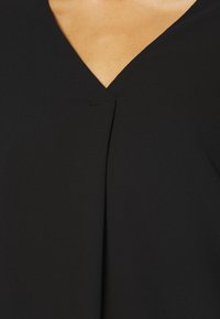 Abbigliamento nero con scollatura a V, caratterizzato da un dettaglio plissettato al centro, realizzato in un tessuto liscio e leggero.