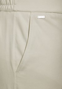 Ljusbeige joggerbyxor gjorda av mjukt, stretchigt tyg med en sidficka och en liten metalletikett. Har ett elastiskt midjeband.