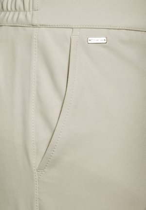 Pantalones jogger de color beige claro, hechos de una tela suave y elástica, con un bolsillo lateral y una pequeña etiqueta de metal. Presenta una cintura elástica.
