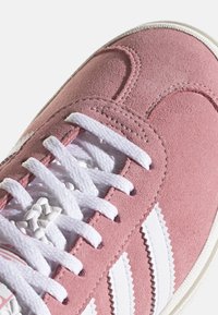 Zapatilla de ante rosa con rayas en zigzag blancas, cordones blancos y una suela blanca texturizada, mostrada en detalle de cerca.
