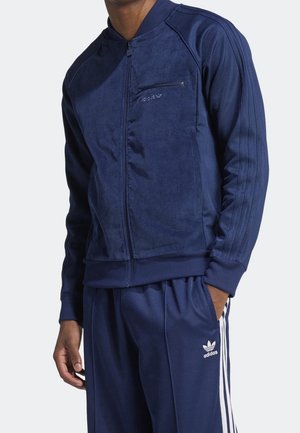 Personne portant un survêtement Adidas bleu marine avec veste zippée et trois bandes blanches sur le pantalon, debout avec une main dans la poche.