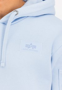 Világoskék kapucnis pulcsi, puha anyagból készült, apró fehér címkével az "Alpha Industries" logójával a mellkasán és fém kiegészítőkkel.