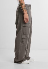 Graue Cargo-Hosen aus Baumwolle mit lockerer Passform. Mehrere Taschen an den Seiten, sichtbare Nähte und eine leichte Verjüngung an den Knöcheln.