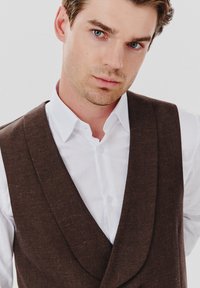 Brązowa kamizelka z teksturowanej tkaniny, z głębokim dekoltem w serek, noszona na białej koszuli. Całość ma dopasowany i skrojony krój.