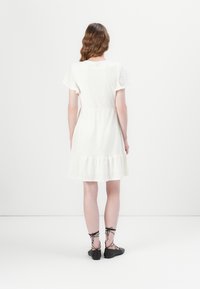 Femme aux cheveux bruns ondulés portant une robe blanche courte en broderie anglaise et des ballerines noires à lacets, debout, de dos, contre un mur blanc uni.