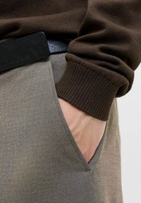 Brown knit sweater with ribbed cuffs, paired with light gray trousers featuring a textured pattern and side pocket. Black belt with embossed branding.