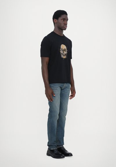 Zwarte t-shirt met schedelafbeelding; korte mouwen, ronde halslijn. Gecombineerd met blauwe spijkerbroek en zwarte leren schoenen.