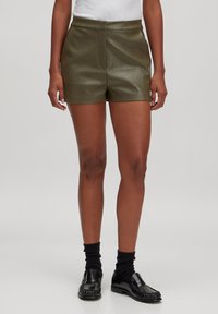 Person trägt dunkelgrüne Leder-Shorts, schwarze Socken und schwarze Loafers und steht vor einem schlichten hellgrauen Hintergrund.