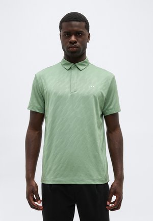 Hombre que lleva una camisa polo de manga corta color verde claro con un patrón texturizado sutil y pantalones negros, de pie contra un fondo liso.