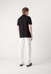 Sort polo skjorte med korte ærmer, parret med hvide bukser. Modellen vender væk, hvilket fremhæver det enkle design og den runde krave. Sorte sko.