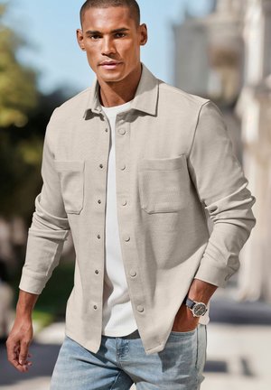 Young man walking outdoors wearing a beige button-up shirt over a white tee, light blue jeans, and a black wristwatch on his left wrist.