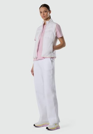 Femme debout portant un gilet blanc sans manches, un chemisier rose à manches courtes, un pantalon blanc et des baskets blanches avec des semelles violettes.