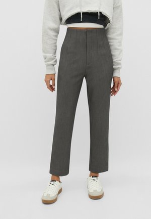 Femme portant un pantalon droit gris taille haute, un sweat à capuche gris clair court et des baskets blanches avec semelles en gomme sur fond uni.