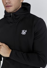 Zwarte rits hoodie met een hoge kraag, gemaakt van rekbaar materiaal. Bevat een geborduurd wit logo op de linkerborst en aansluitende mouwen.