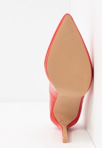 Chaussure à talon haut rouge vue par la semelle, bout pointu, talon fin, placée contre une surface blanche et un coin de mur.