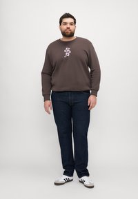 Brown sweatshirt with floral embroidery in white and pink, paired with dark blue jeans and white sneakers with black stripes.