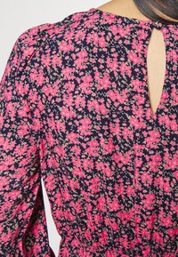 Gros plan d'une robe bleu marine à motif floral rose, avec une découpe goutte d'eau dans le dos et un tissu froncé à la taille et aux épaules.
