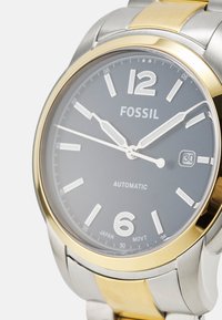 Montre en acier inoxydable argent et or avec un cadran gris foncé, comportant de grands chiffres blancs, un mouvement automatique et un guichet de date à 3 heures.